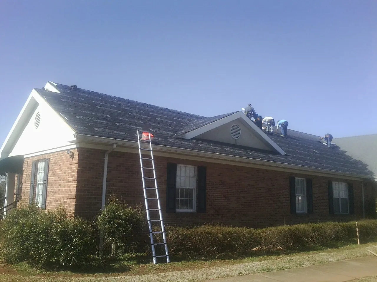 Crew tearing shingles off a brick ranch roof with a ladder set — Emergency Roof Repair work by our crew in Drake