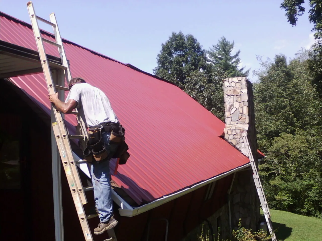 Roofer on a ladder beside a red metal roof and stone chimney — Soffit & Fascia Repair by Roofing Brothers in Drake